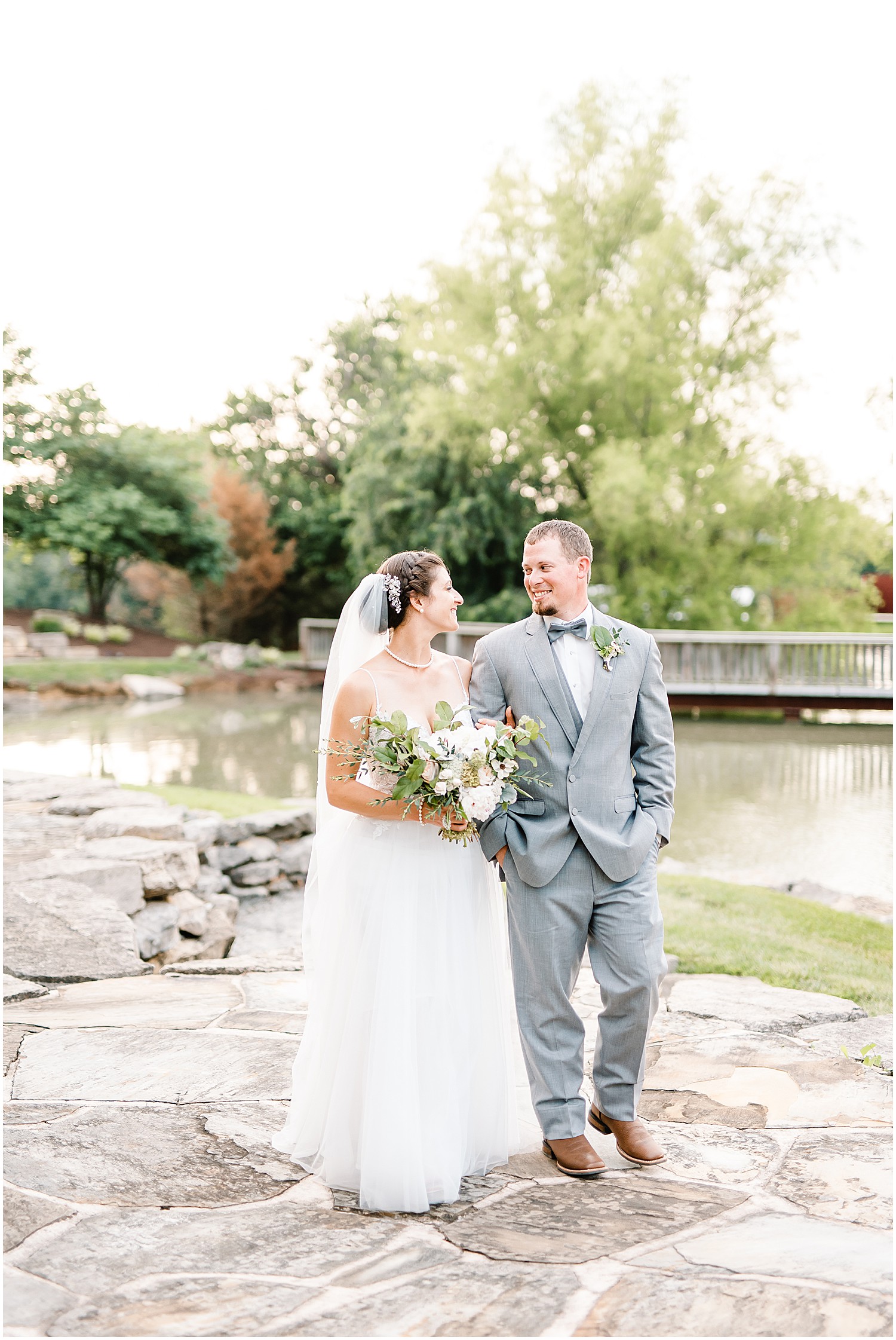 a bride and groom walk on the rock patio by the lake and bridge at cedar lake cellars during a July wedding