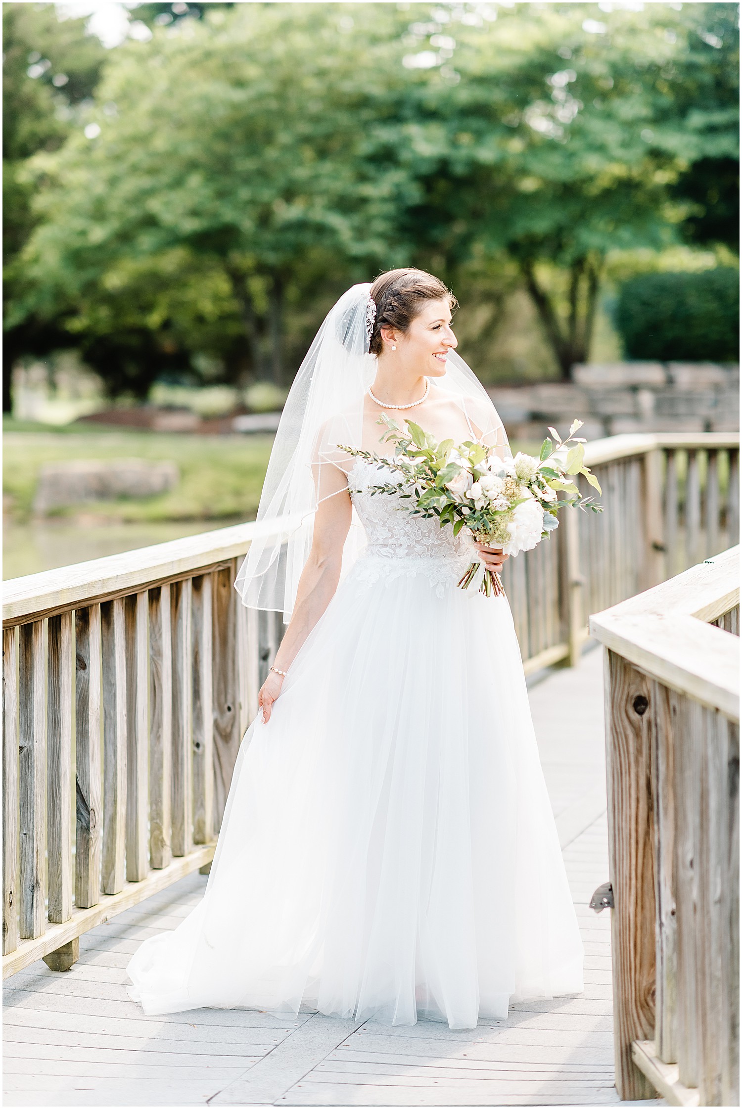 bride in veil stands on bridge looking out over lake on her wedding day at cedar lake cellars