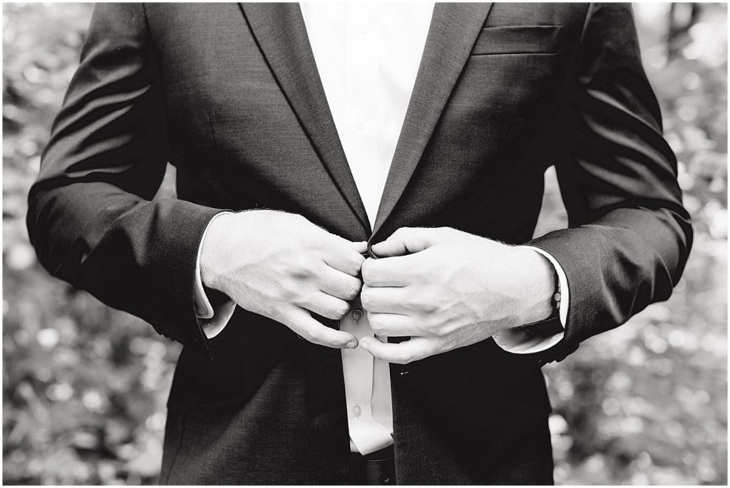 back and white image of groom buttoning jacket before wedding