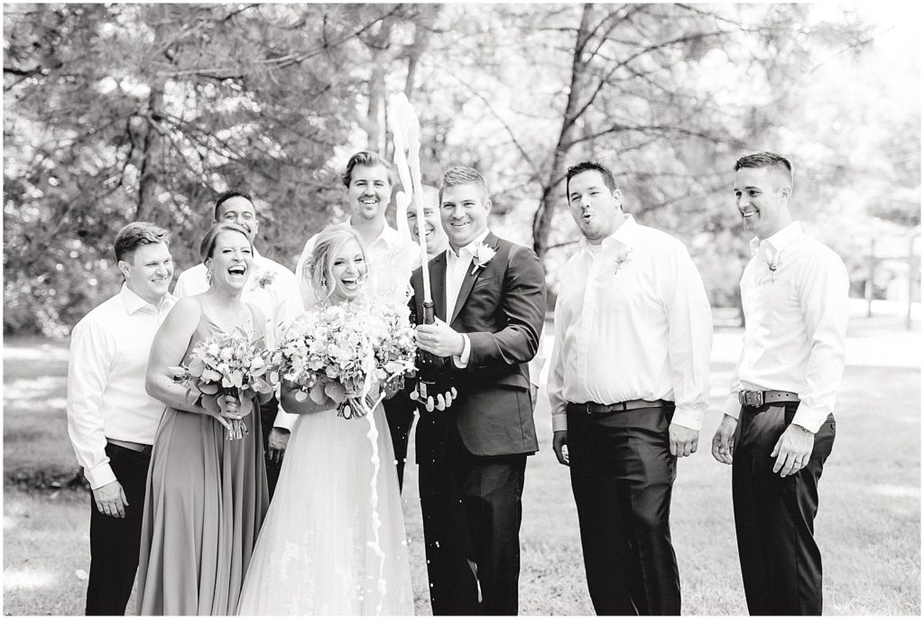 black and white image of groom popping champagne surrounded by wedding party during wedding day
