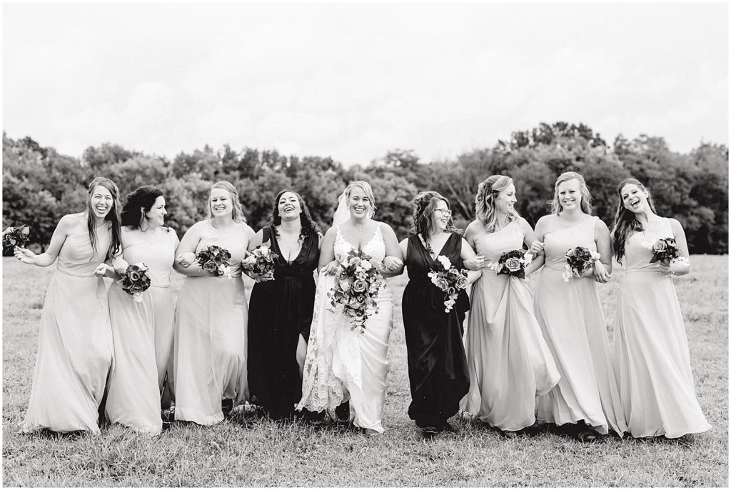 black and white image of bride and bridesmaids walking through field arm in arm