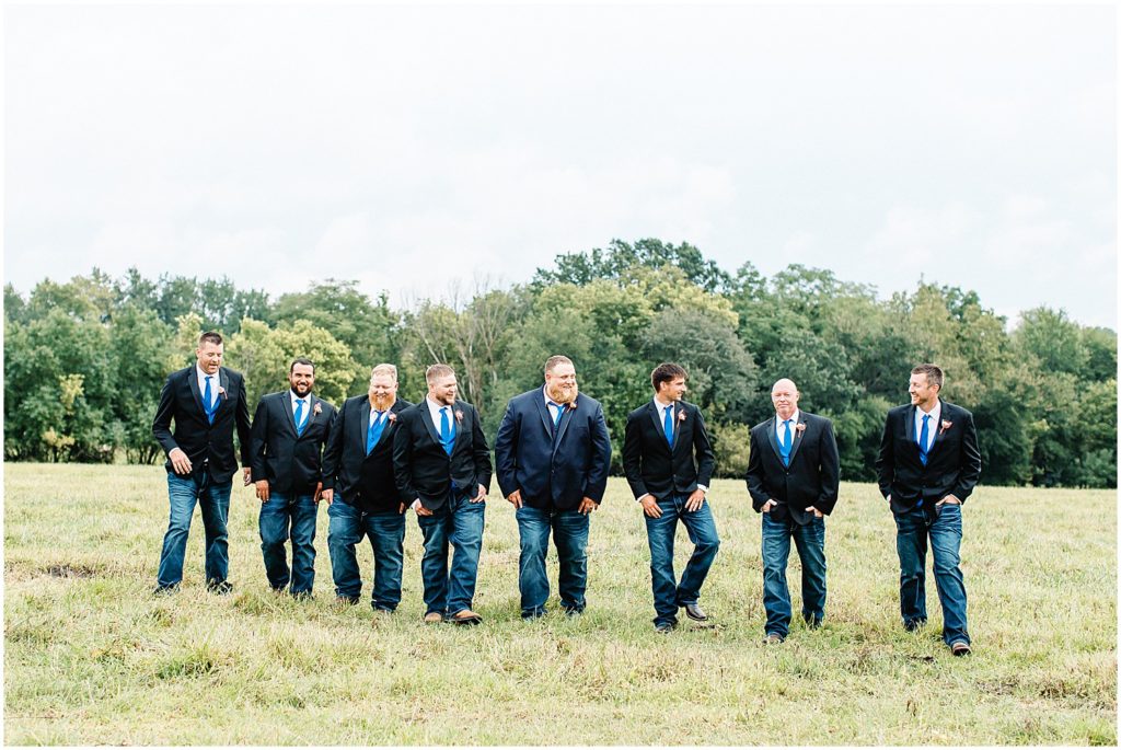 groom and groomsmen wearing jackets jeans and boots walking in field during wedding party portraits