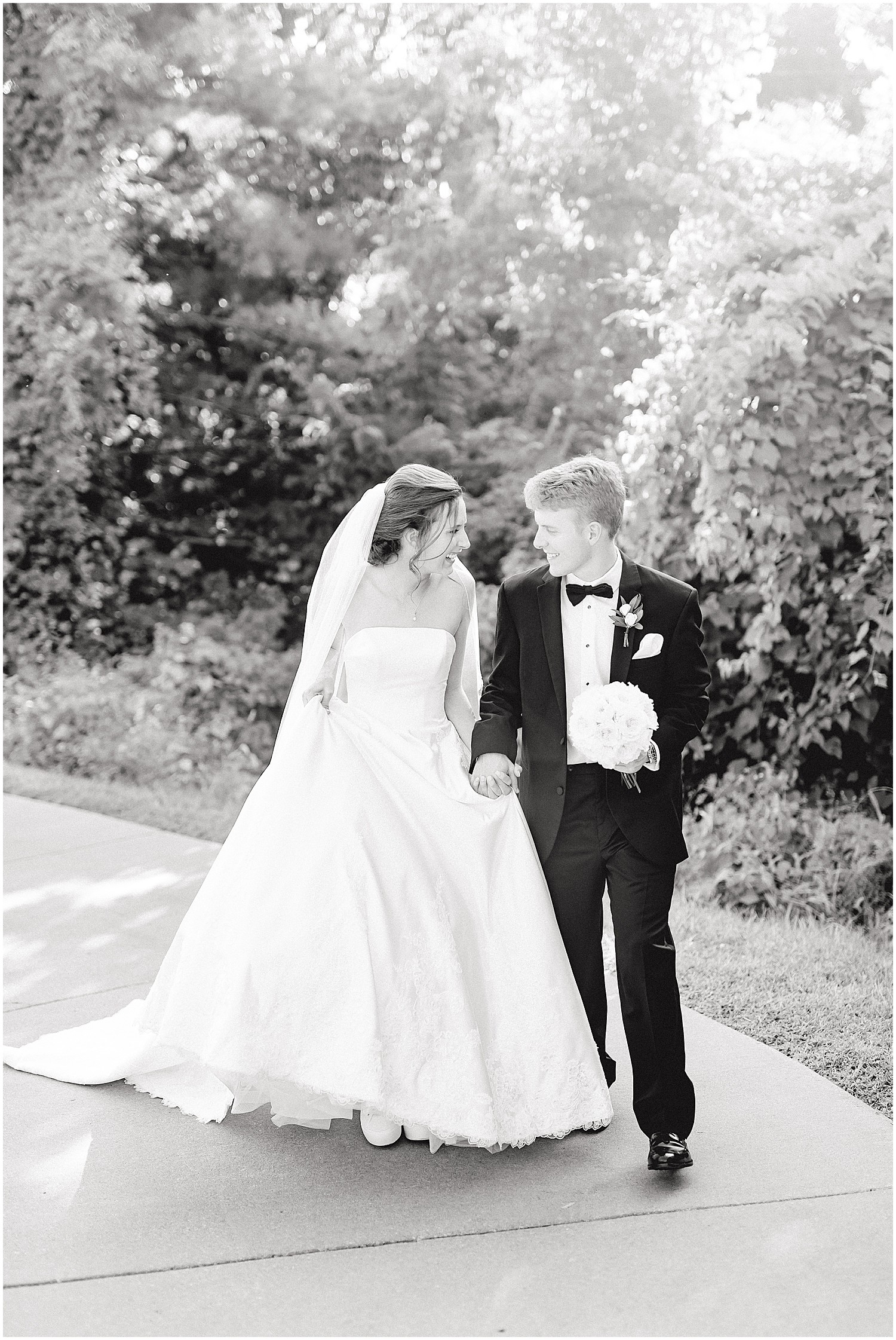 black and white image of bride and groom smiling and walking on sidewalk during portraits on wedding day