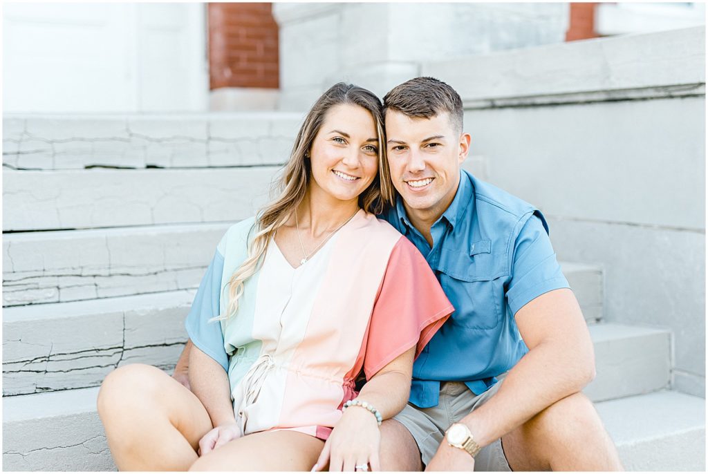 engaged couple smiles at camera sitting on concrete stairs outside gasconade county courthouse