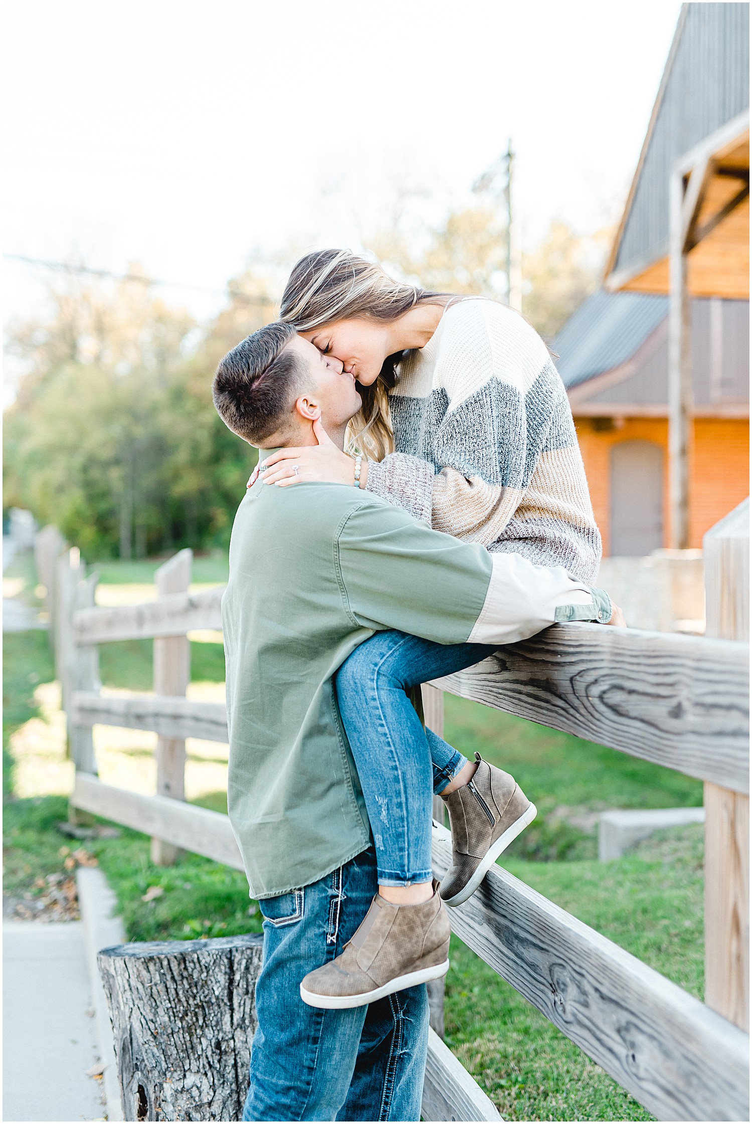 girl sits on fence while man kisses her during Hermann Farm engagement session