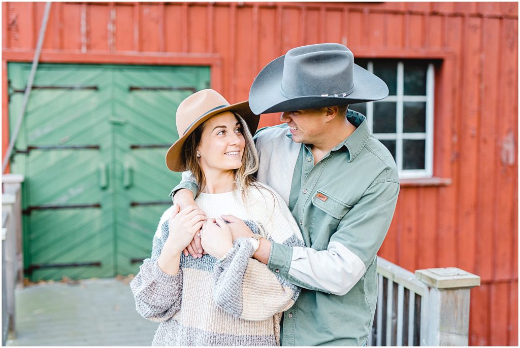 couple smiles at each other wearing cowboy hats in front of green door during engagement session