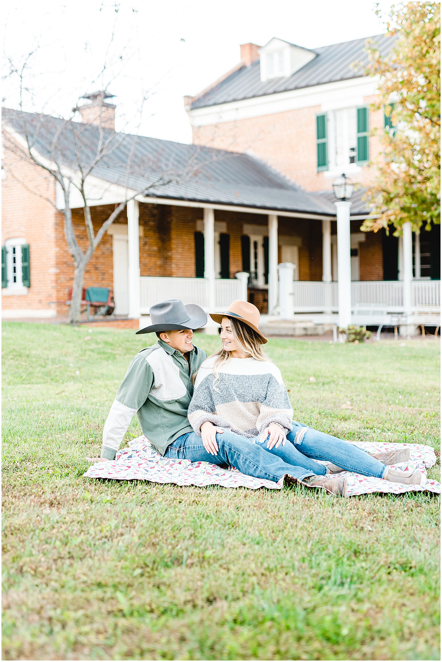 couple wearing cowboy hats smile at each other while sitting on blanket in grass during Hermann, mo engagement session