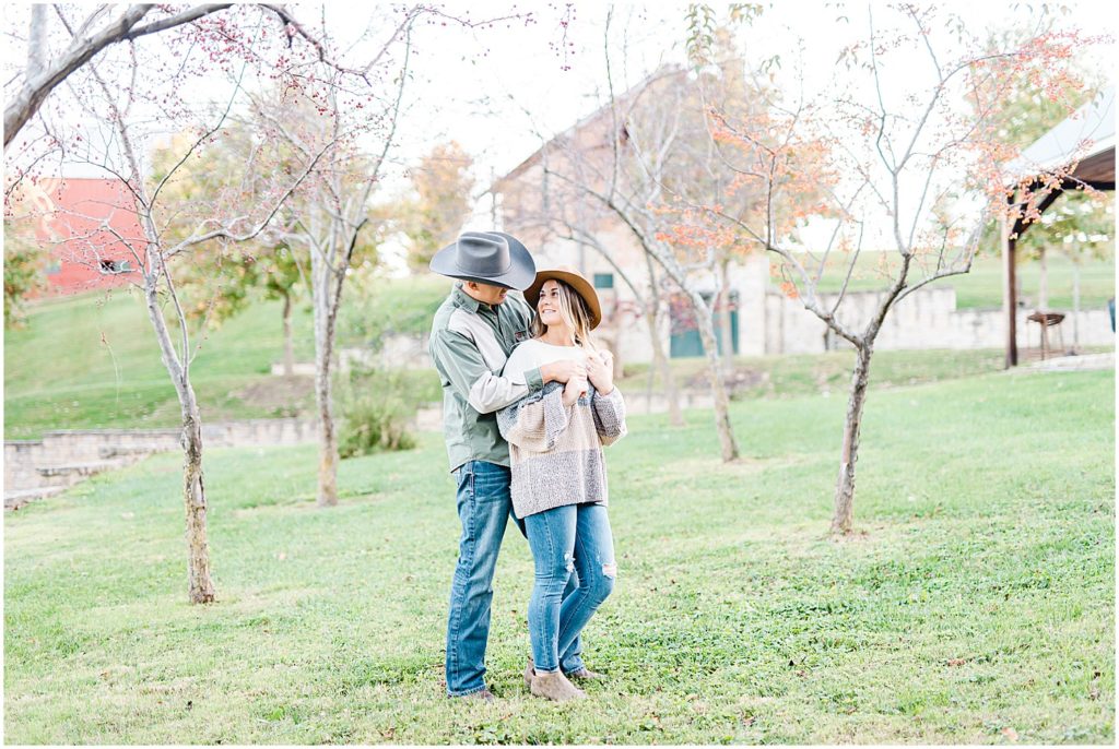engaged couple smile at each other in tree grove during Hermann, mo engagement photos