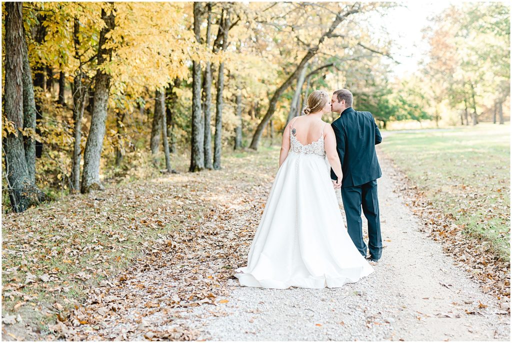 bride and groom kiss while their backs are turned on tree lined gravel lane during pictures on wedding day