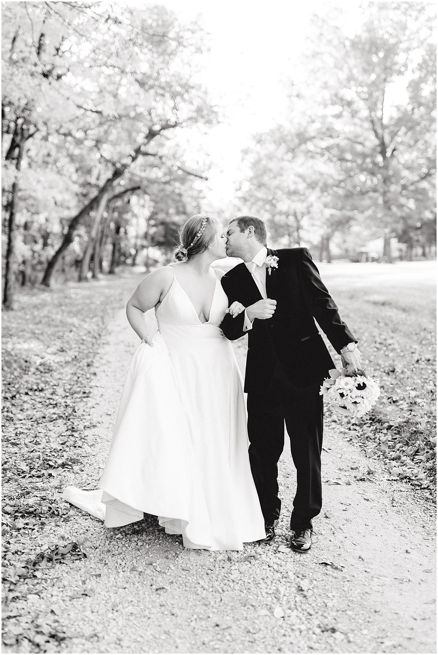 black and white image of bride and groom kissing while they walk on gravel drive under trees on wedding day