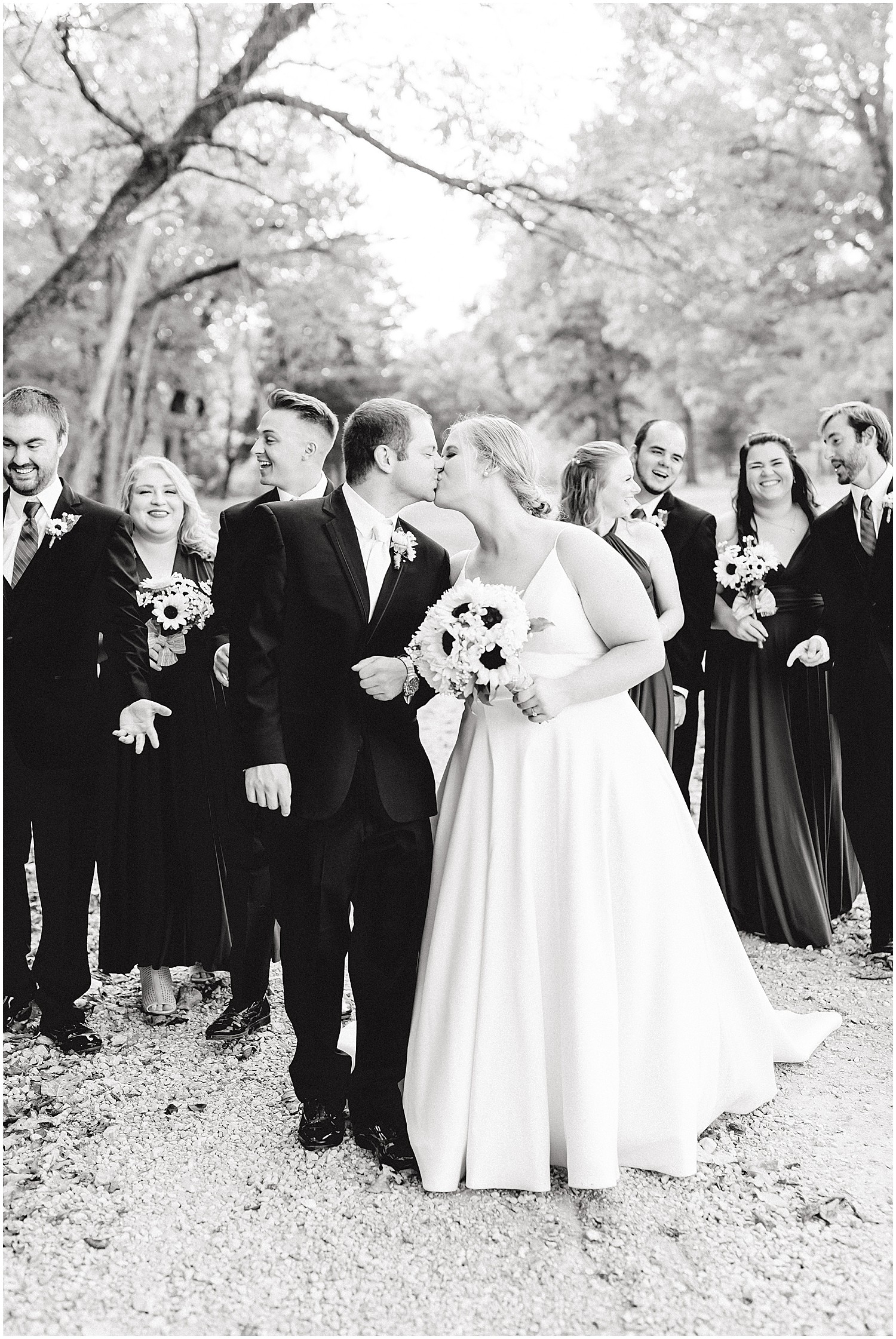 black and white image of bride and groom kissing while wedding party walks behind them