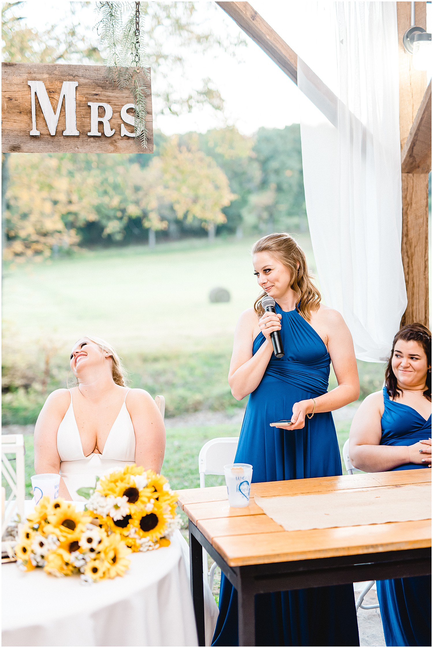 bride and maid of honor laugh during speeches at wedding reception at kempker's back 40