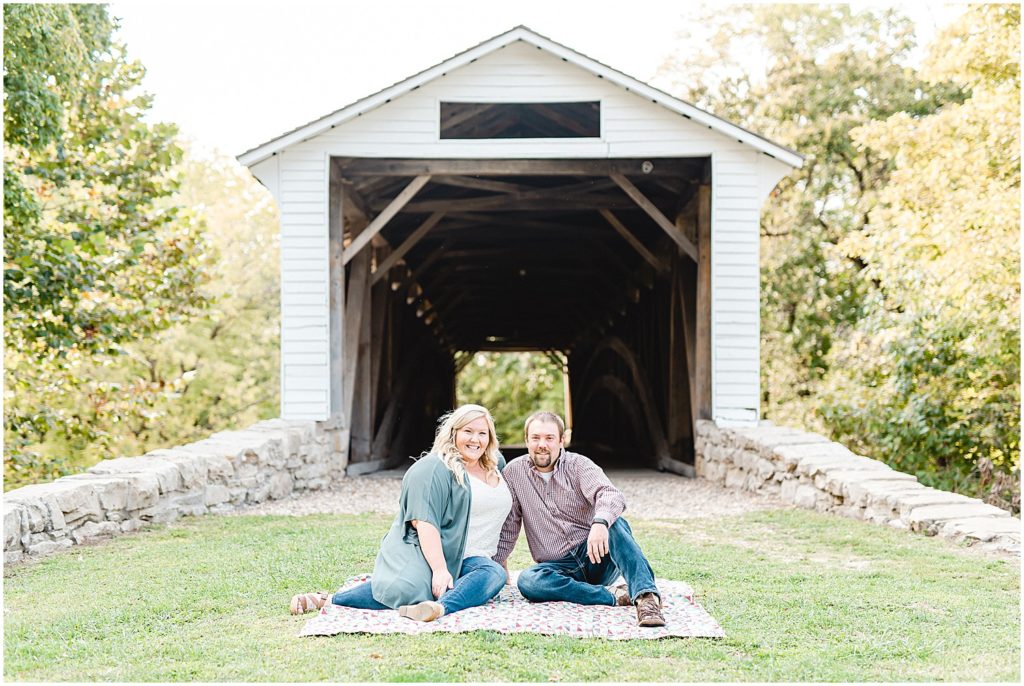 couple sitting on blanket in front of white covered bridge for engagement session