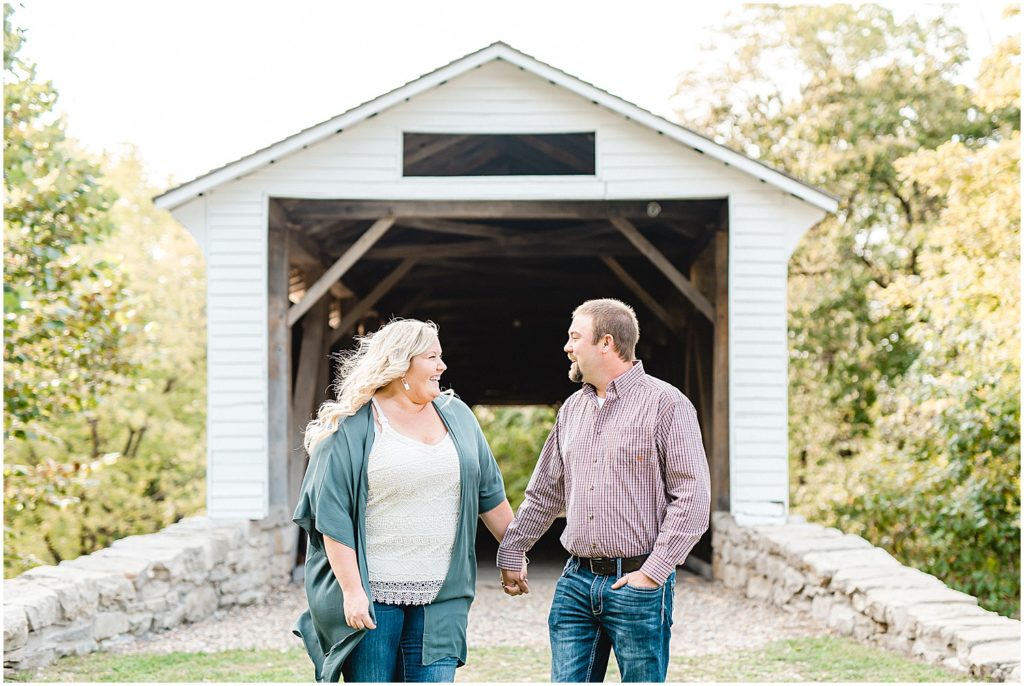 Couple smiles at each other in front of white covered bridge in Missouri for engagement session