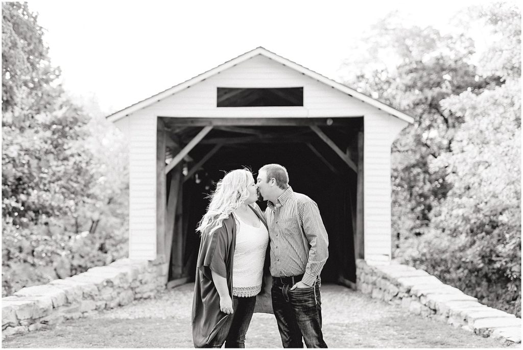 Black and white image of couple kissing in front of bridge during engagement pictures