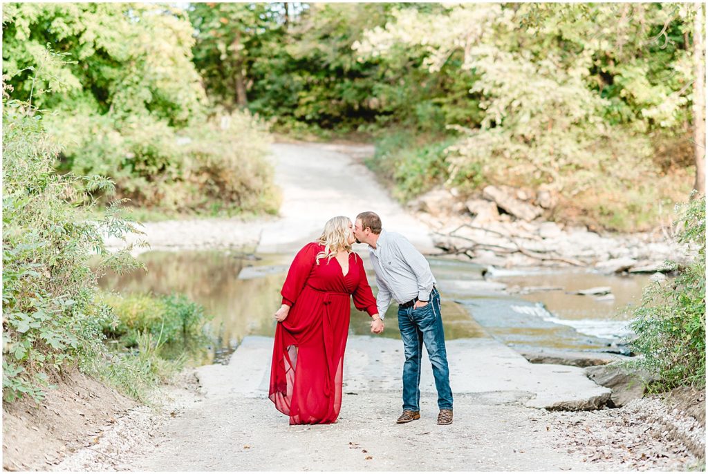 Couple kisses on road in front of creek wearing long maroon dress for engagement pictures.