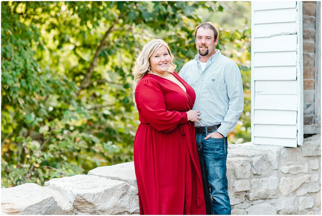 Couple smiles at the camera in front of rock wall during engagement session wearing long maroon dress.