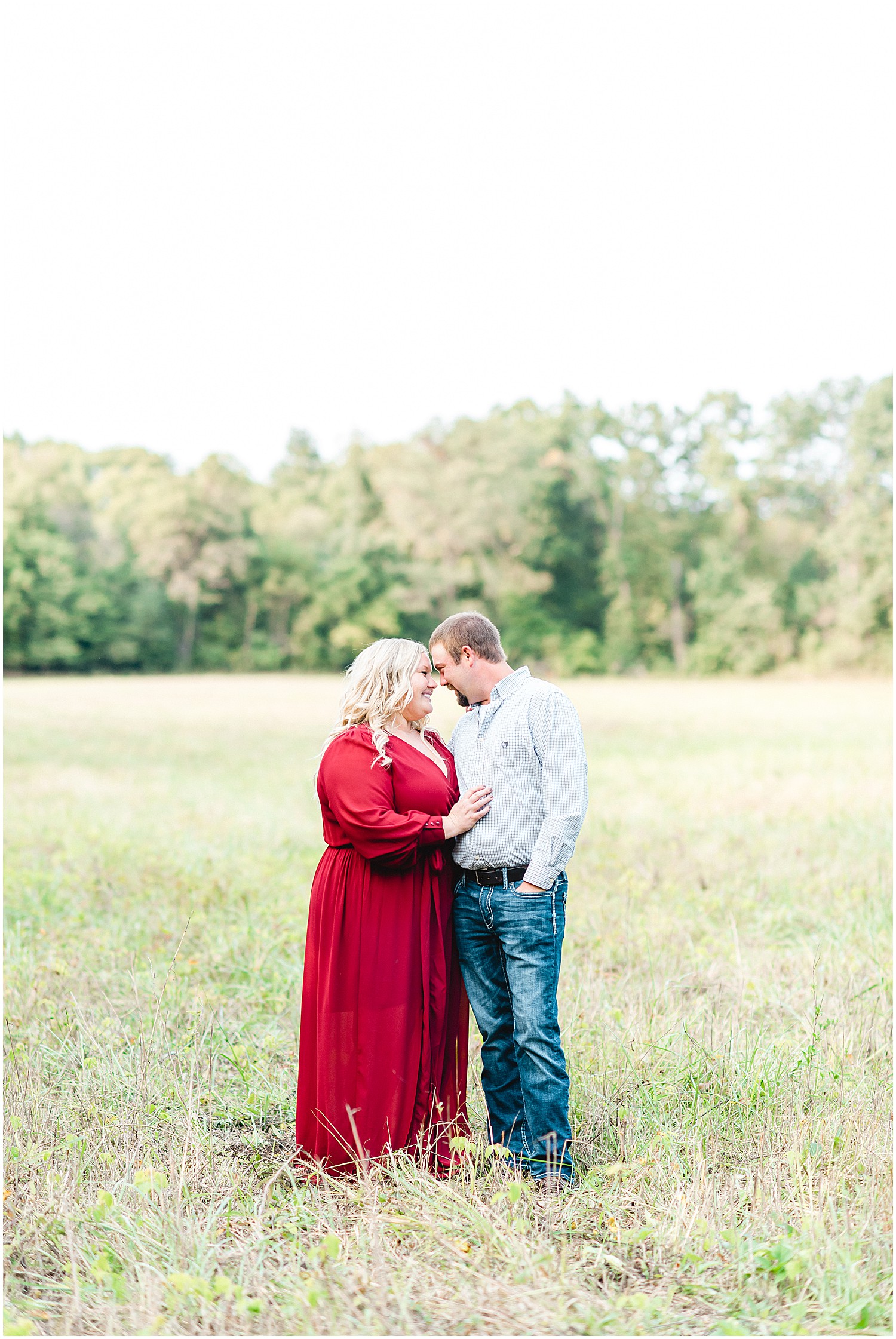 Couple stands in field smiling during engagement session wearing maroon dress.