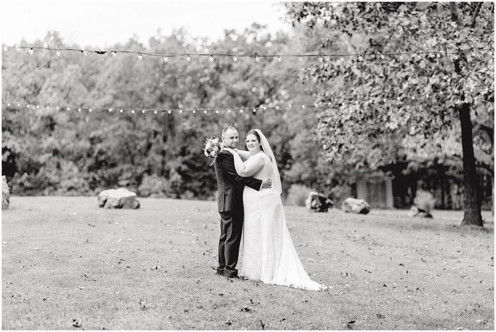 black and white image of bride and groom hugging in grass under patio lights at wildflower wedding venue