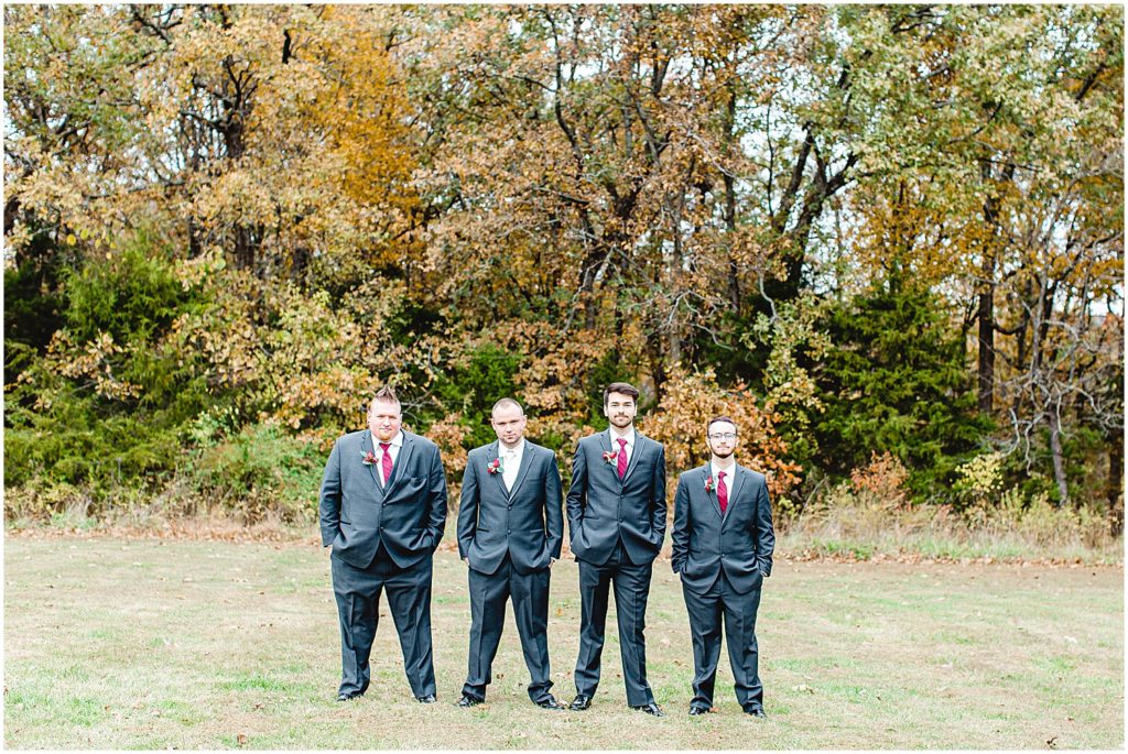 groom and groomsmen in dark gray suits and maroon ties stand in a line during wedding party portraits 