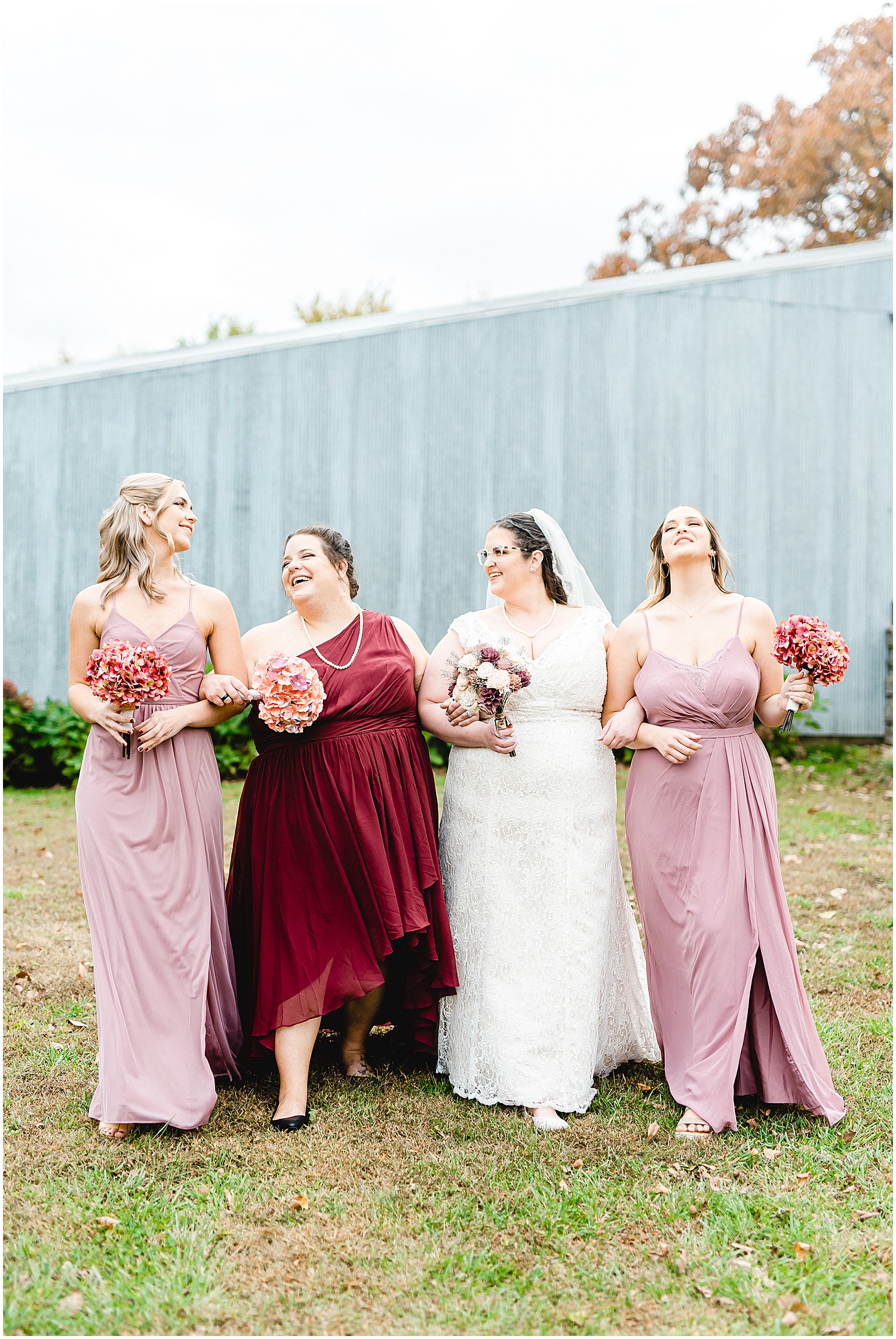 bride and bridesmaids wearing shades of pink and maroon smile as they walk during portraits on wedding day at wildflower in Eugene, mo