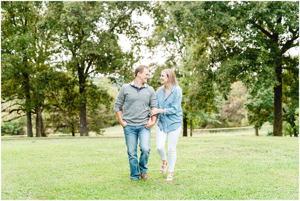 couple walks hand in hand on grass smiling at each other for Missouri farm engagement session