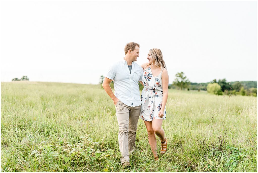 couple walks through grassy field for engagement pictures in missouri