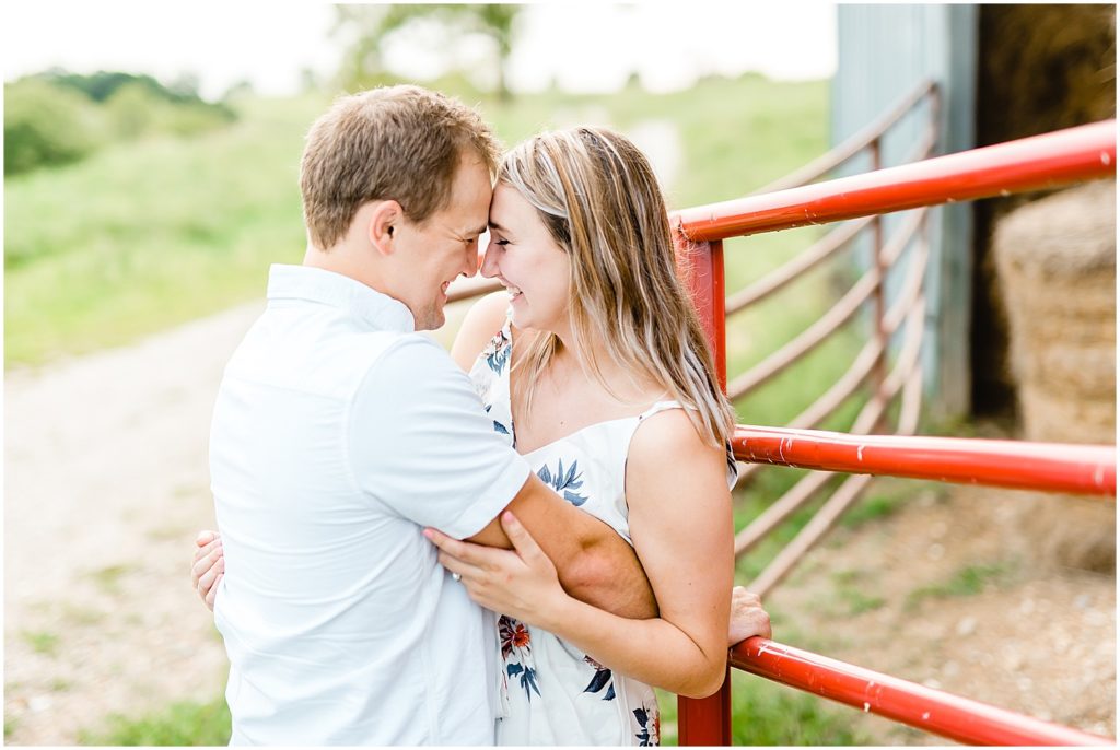 couple smiles against red fence with hay in the background for engagement session on farm