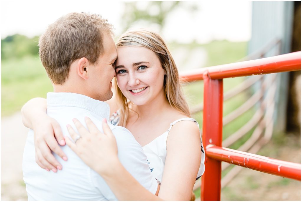 woman smiles at camera while hugging fiance during engagement pictures on family farm in westphalia, mo