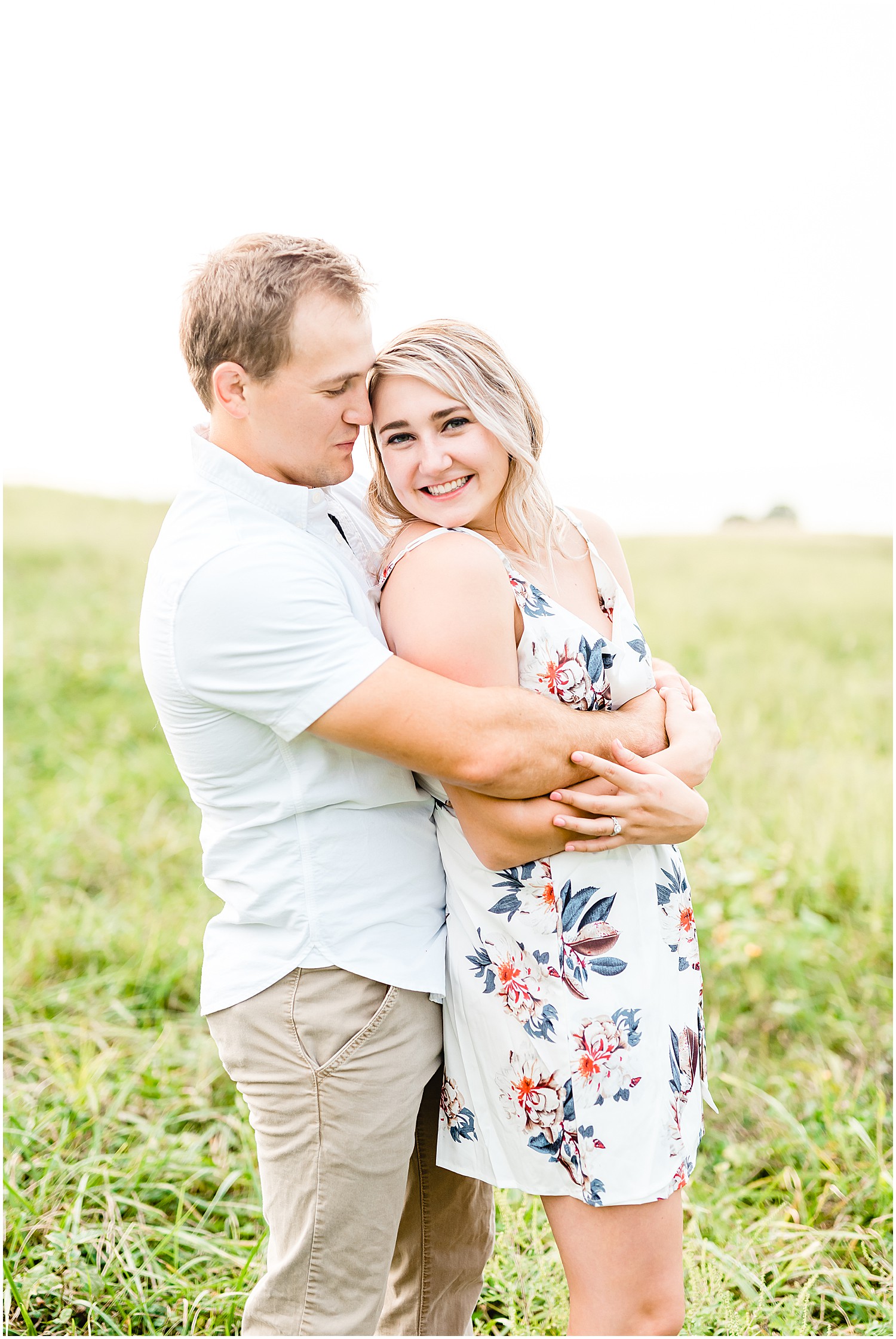 couple hugs and smiles at the camera standing in the grass on farm for engagement pictures
