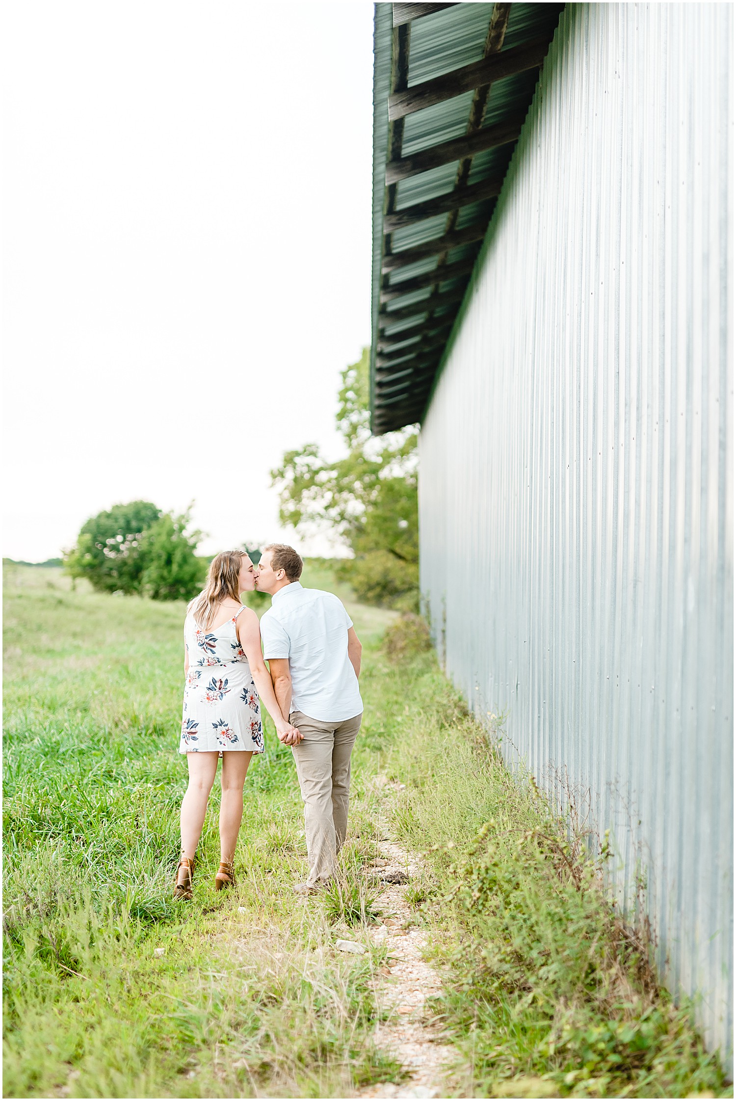 couple kisses and walks next to gray metal barn during engagement session on missouri farm