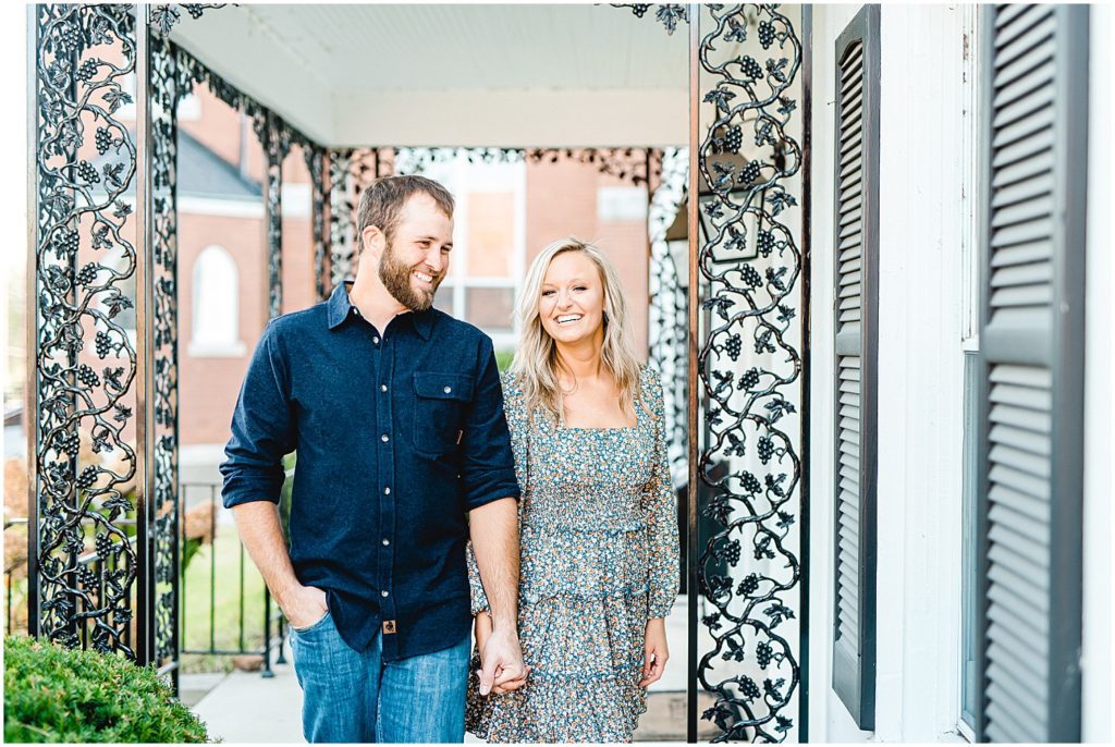 couple smiling and walking by White House with black shutters during missouri engagement session