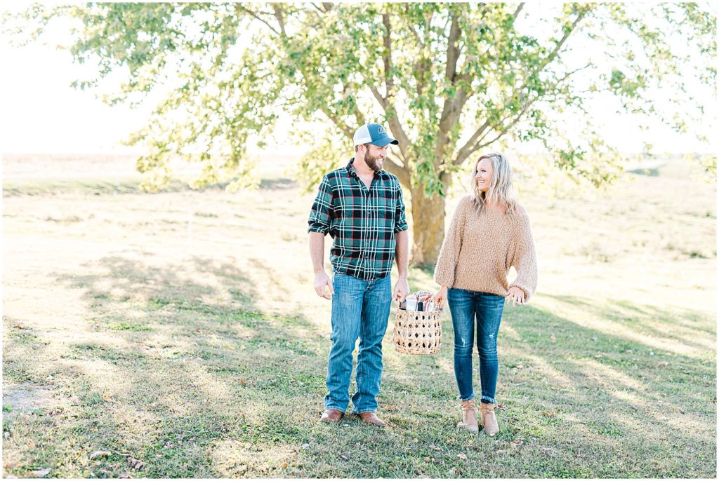 couple walks together under tree holding picnic basket for engagement session