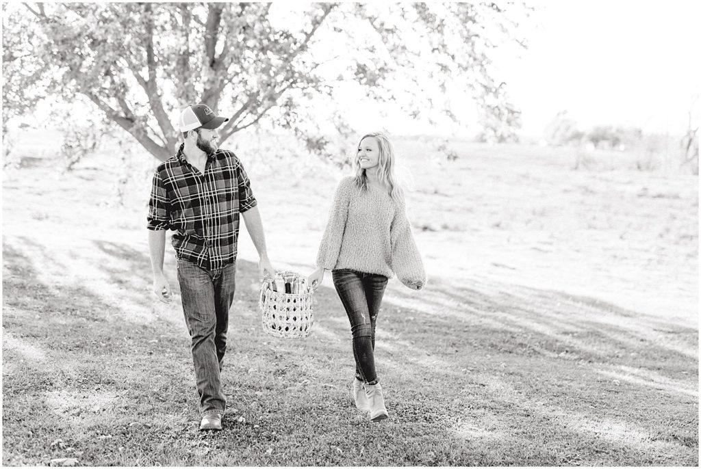 black and white image of couple walking with picnic basket under tree for engagement pictures in missouri