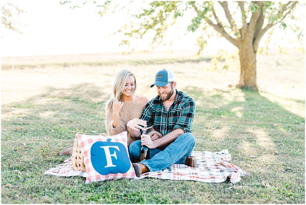 man opens wine during engagement session on picnic blanket