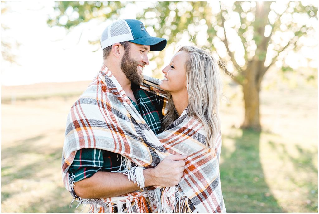 young couple smiles at each other wrapped in blanket during engagement session
