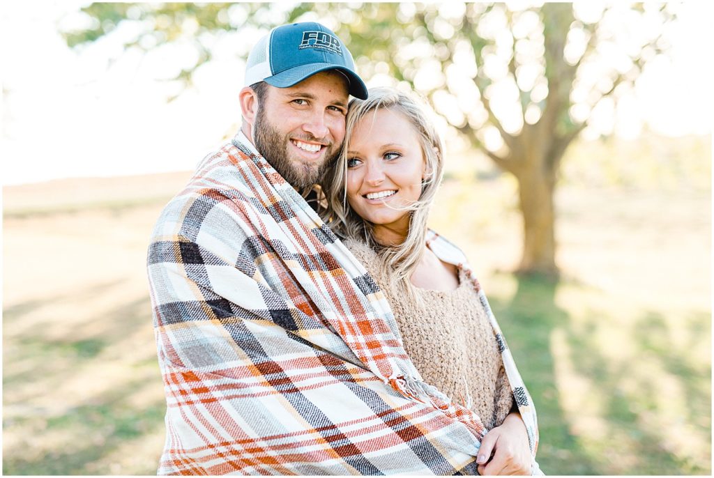 couple snuggles together wrapped in plaid blanket for engagement pictures