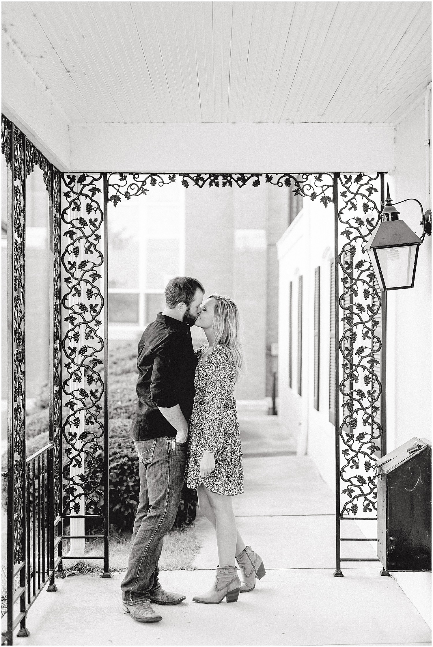 black and white image of couple kissing under porch with black iron details during engagement session in missouri