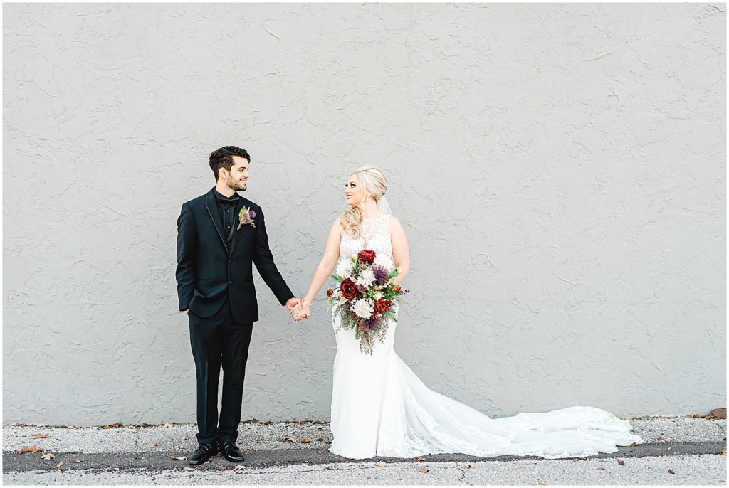 bride and groom hold hands and look at each other while standing against gray wall