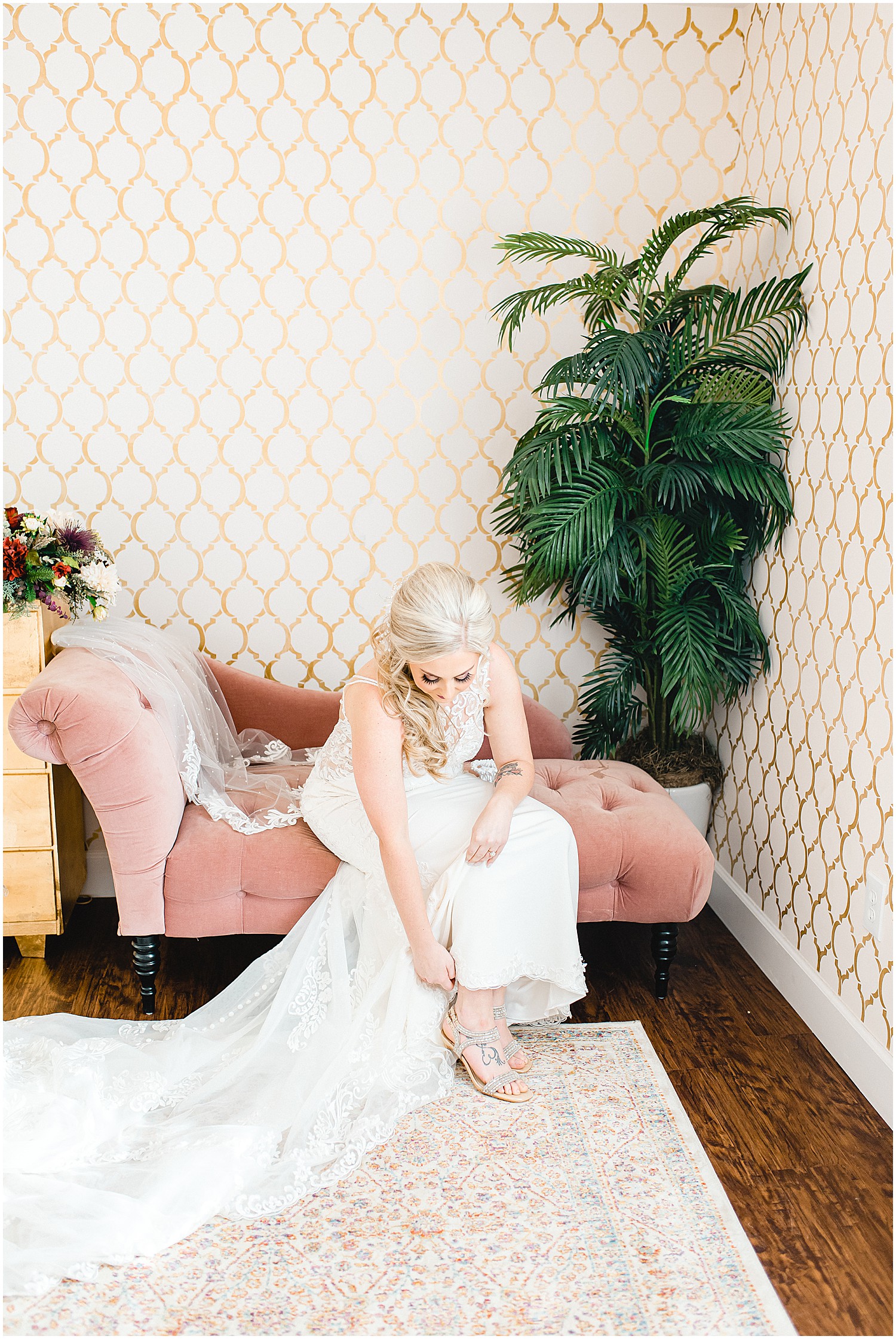 bride sits on pink sofa while putting her wedding day shoes on