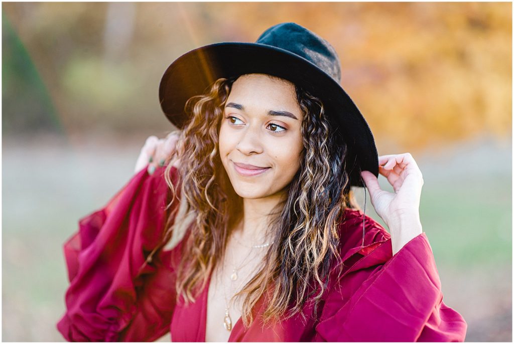 senior girl stands on grass smiling at camera with yellow tree in background wearing maroon dress and black hat for senior pictures