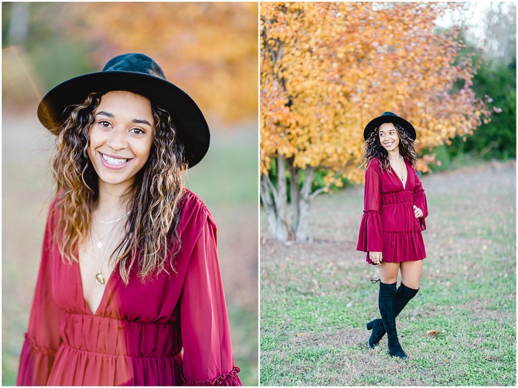 senior girl stands on grass smiling at camera with yellow tree in background wearing maroon dress and black hat for senior pictures
