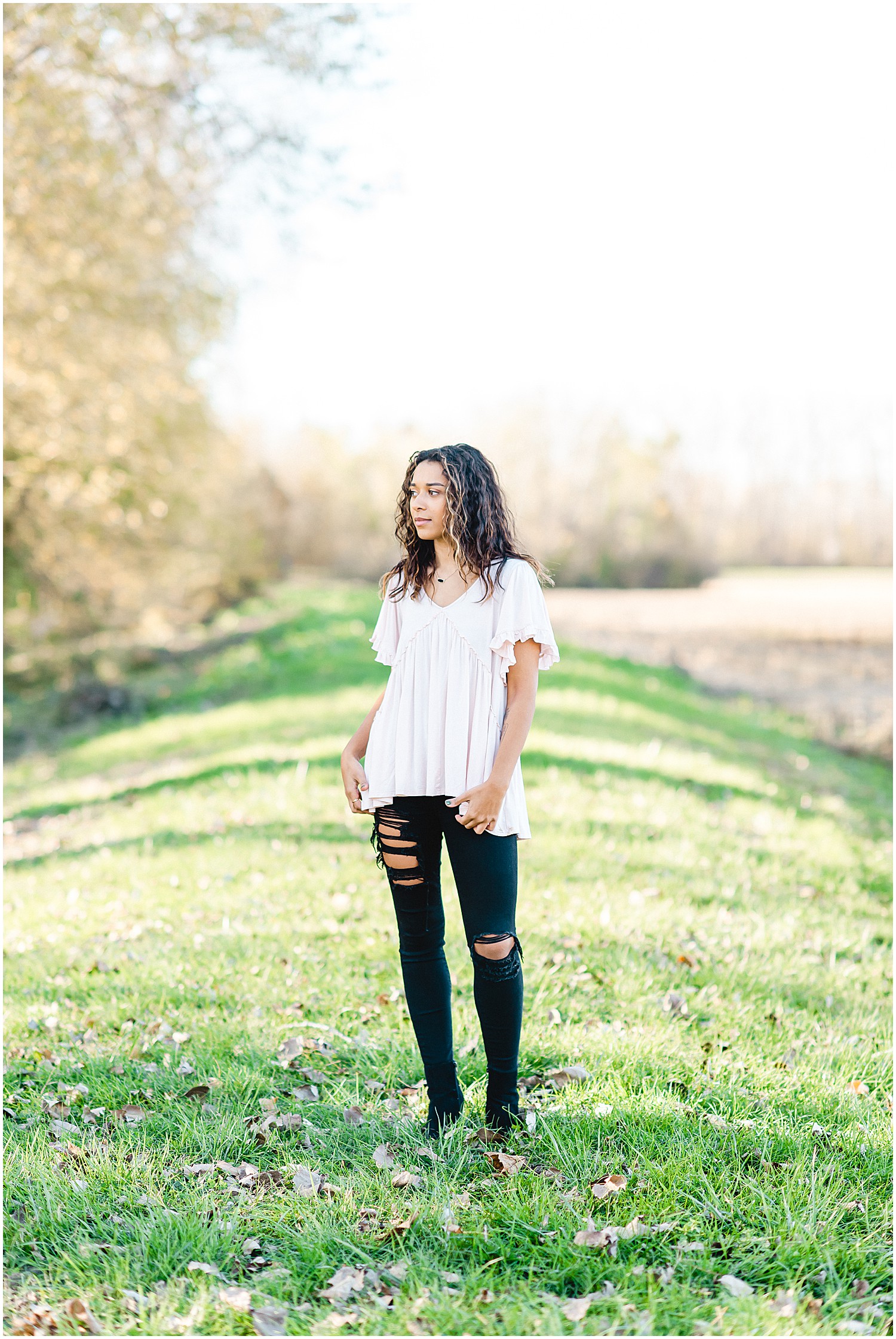 Senior girl wearing ripped black skinny jeans standing on grass staring off to the side during senior pictures