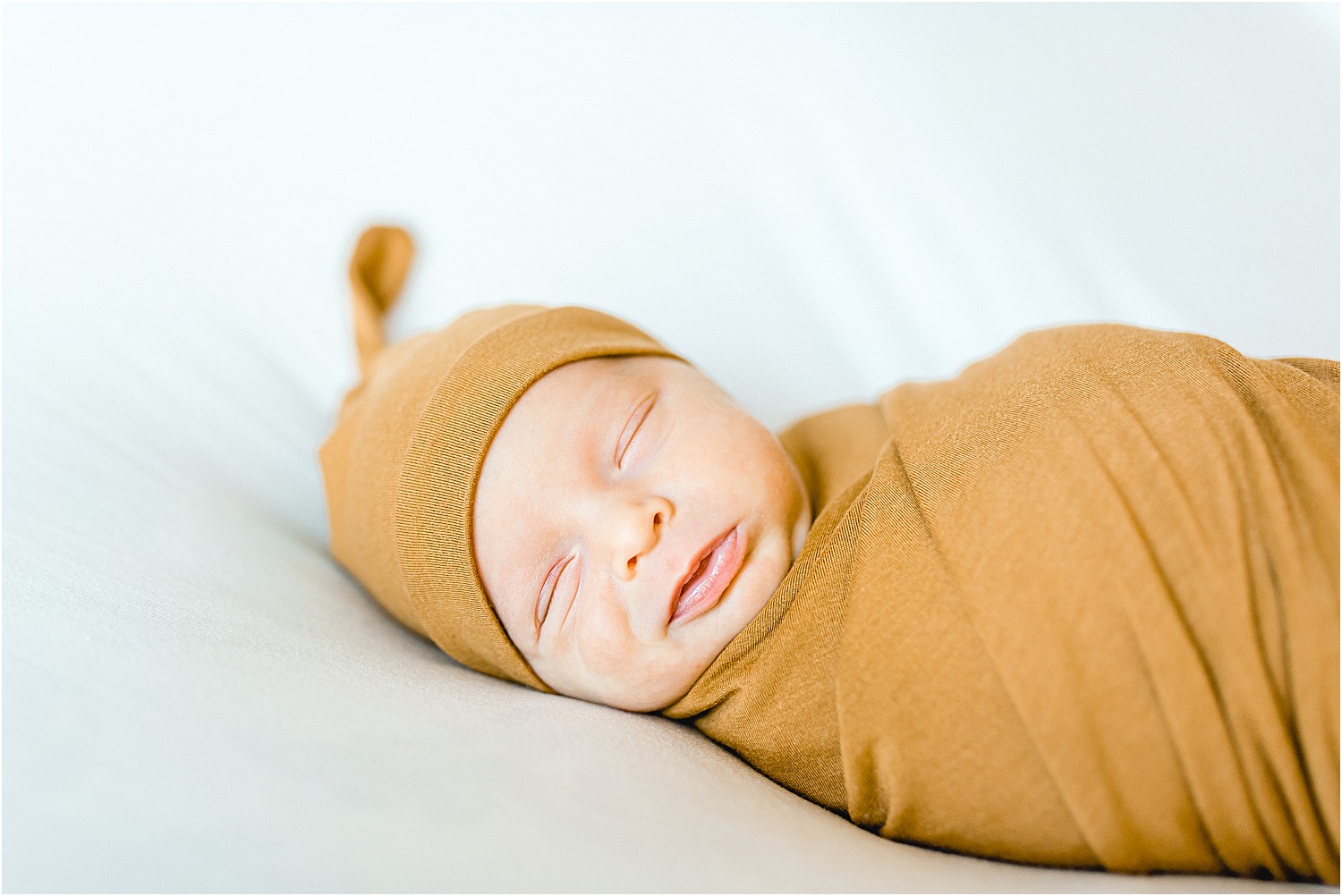 newborn baby boy in gold swaddle and hat lays on white blanket