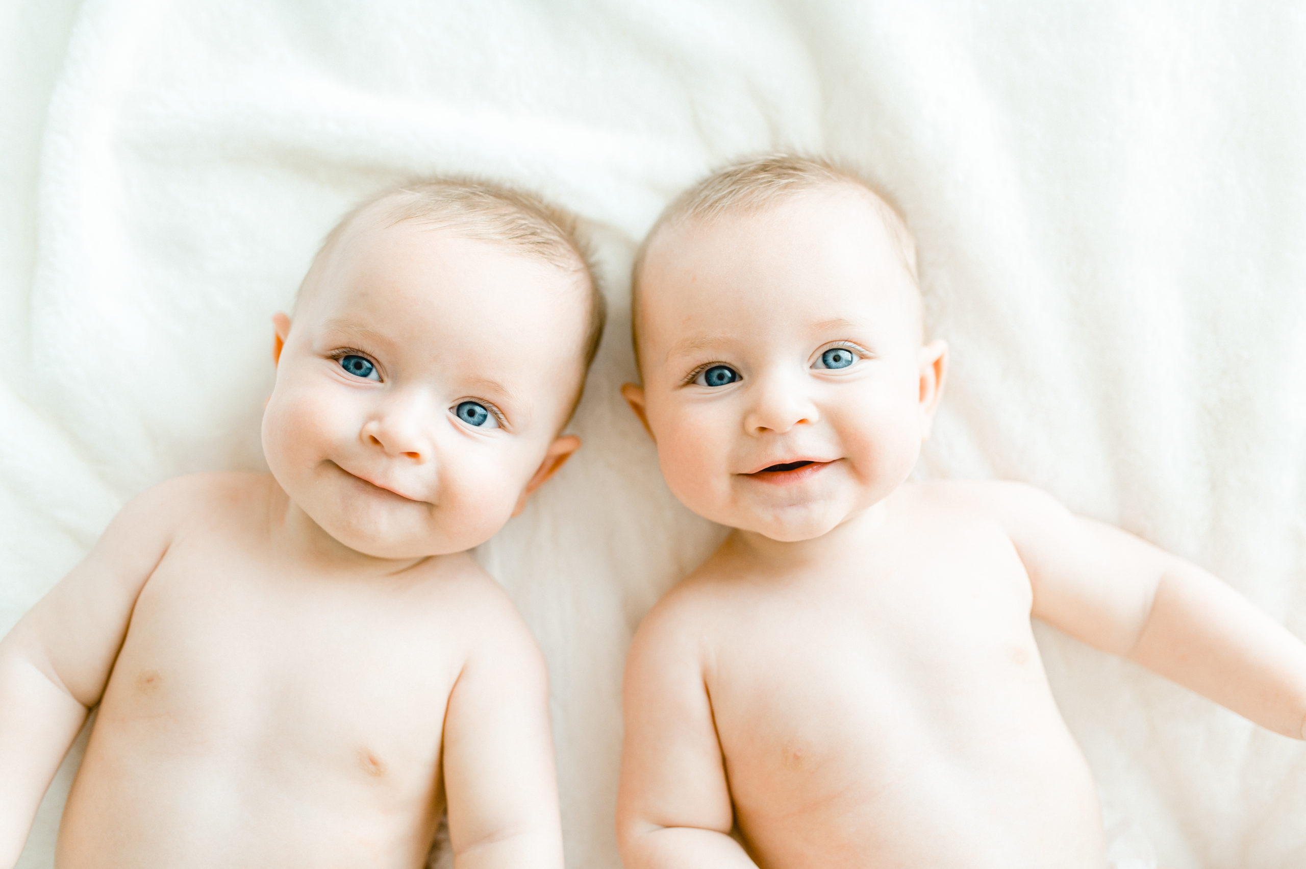 Twin 6 month old boys lay on white blanket smiling